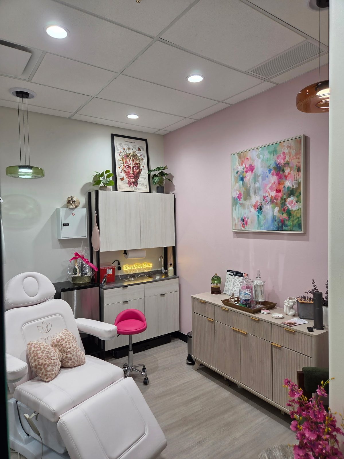 Treatment room with a white treatment chair, a pink accent wall and a floral painting at Infinite Glow